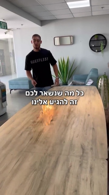 גבר צעיר בחולצה שחורה עומד ליד שולחן עץ עם להבה באולם רהיטים, קאבר רילס עם הכיתוב "כל מה שנשאר לכם זה להגיע אלינו"